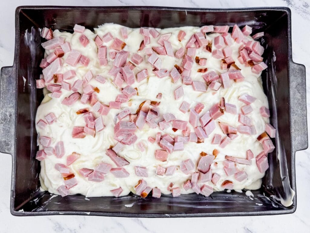 A baking pan filled with uncooked dough mixed with white and pink diced ingredients, likely ham or similar, on a marble countertop.