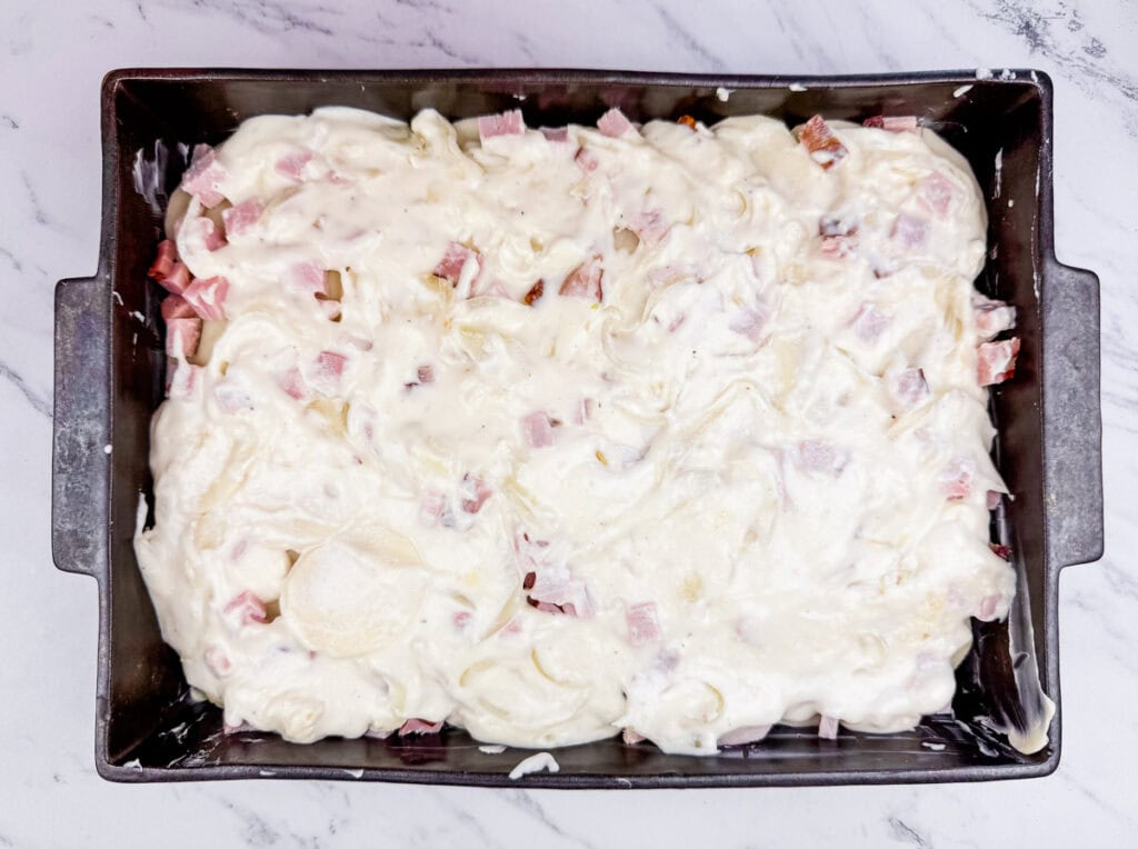 Baking pan with uncooked creamy pasta bake, containing white sauce, diced ham, and pasta, on a marble countertop.
