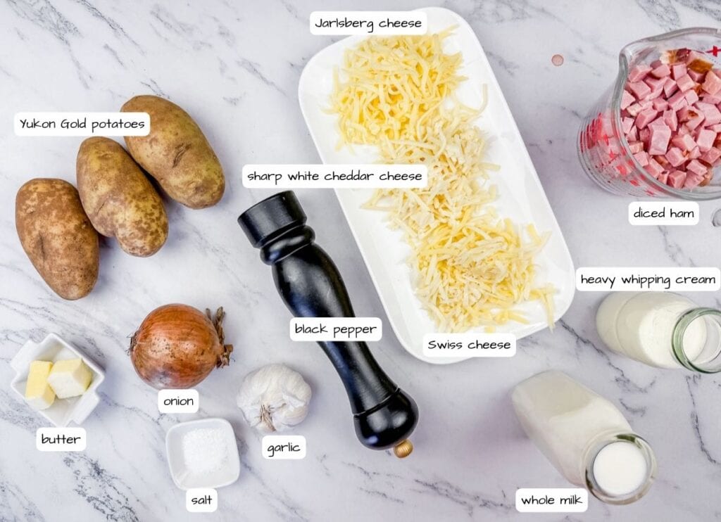 Yukon gold potatoes, cheeses, diced ham, an onion, garlic, butter, salt, black pepper, heavy cream, and whole milk on a marble surface.