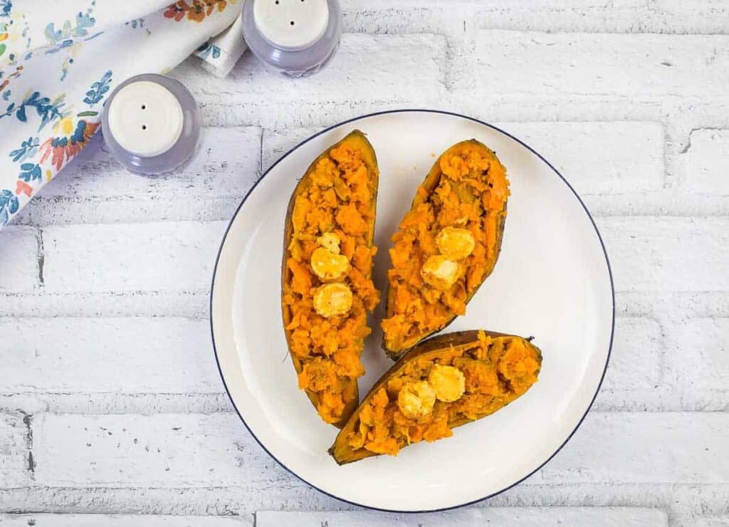 Three halves of roasted sweet potatoes topped with butter on a white plate, set on a white brick surface with a floral napkin and salt and pepper shakers nearby.