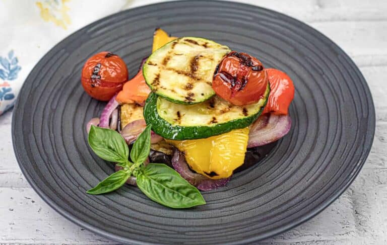 Grilled zucchini, tomatoes, red onions, and bell peppers are stacked on a black plate, garnished with fresh basil leaves.