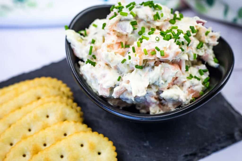 A black bowl of creamy salmon dip topped with chopped chives is placed on a slate board next to a row of round crackers.