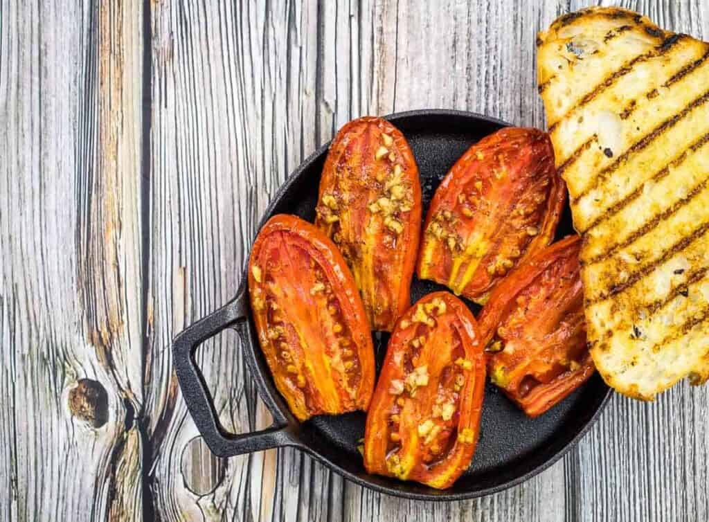 Grilled tomato halves with garlic and spices in a cast iron skillet next to slices of grilled bread on a wooden surface.