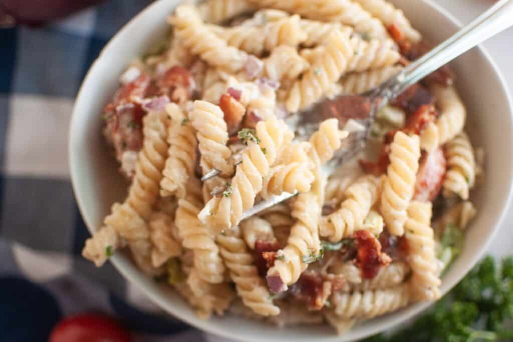 A fork lifts a portion of BLT pasta salad with spiral pasta, creamy dressing, and pieces of bacon from a white bowl. Red onions and herbs are also visible in the salad. A blue checkered cloth and cherry tomatoes are in the background.