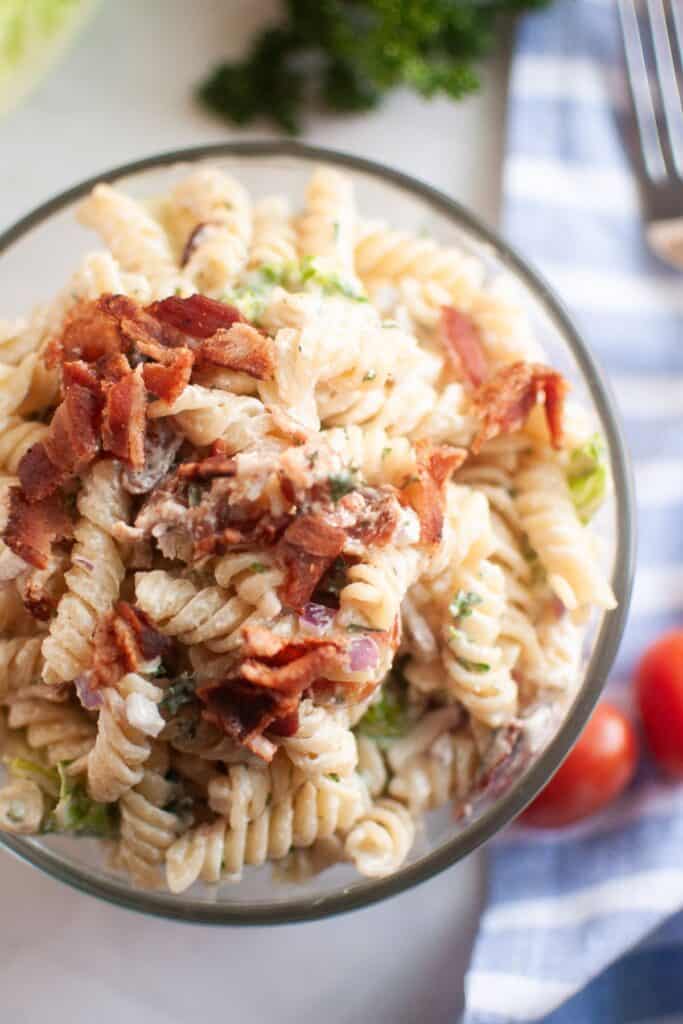 Overhead shot of a glass bowl filled with BLT pasta salad, featuring spiral pasta, creamy dressing, pieces of bacon, and other visible ingredients. A blue striped cloth and cherry tomatoes are visible in the background.