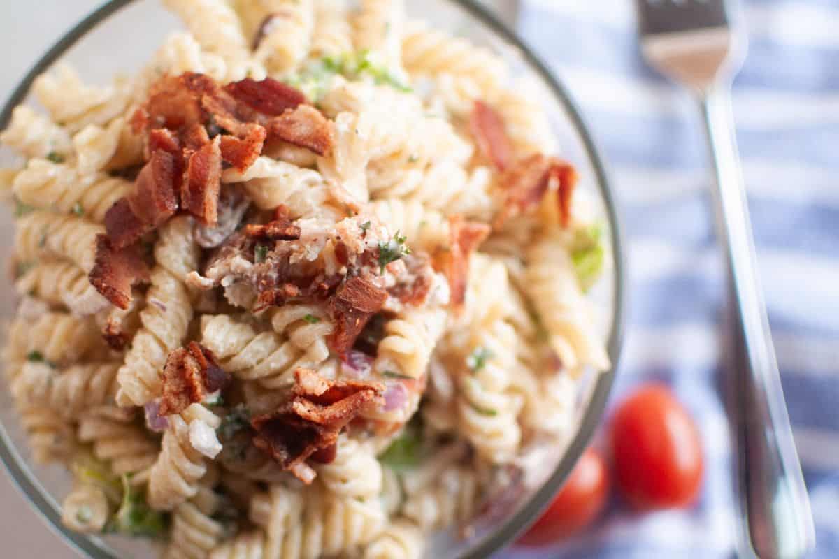 Overhead shot of a glass bowl filled with pasta salad, featuring spiral pasta, creamy dressing, pieces of bacon, and other visible ingredients. A blue striped cloth and cherry tomatoes are visible in the background.