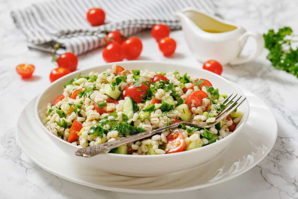 A bowl of barley salad with chopped tomatoes, cucumbers, parsley, and green onions, served with a fork. Olive oil and fresh cherry tomatoes are in the background.