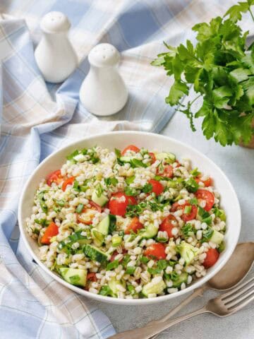 A bowl of barley salad with chopped cucumber, cherry tomatoes, herbs, and green onions on a table with a blue plaid cloth, parsley, and salt and pepper shakers.