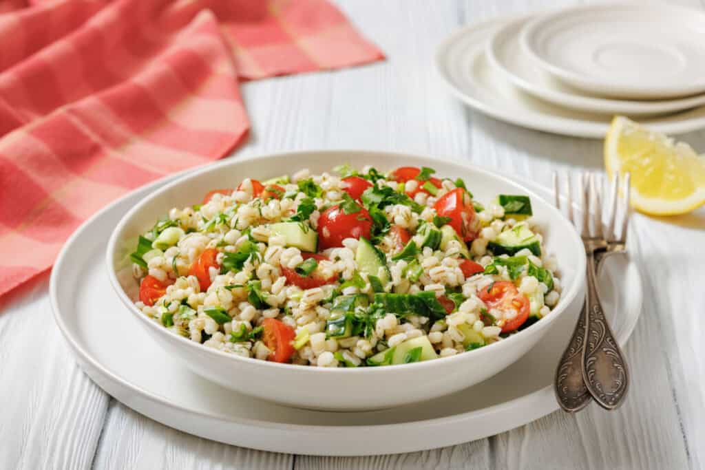 A white bowl of barley salad with tomatoes, cucumbers, and herbs sits on a plate next to utensils, with a lemon wedge and napkin in the background.
