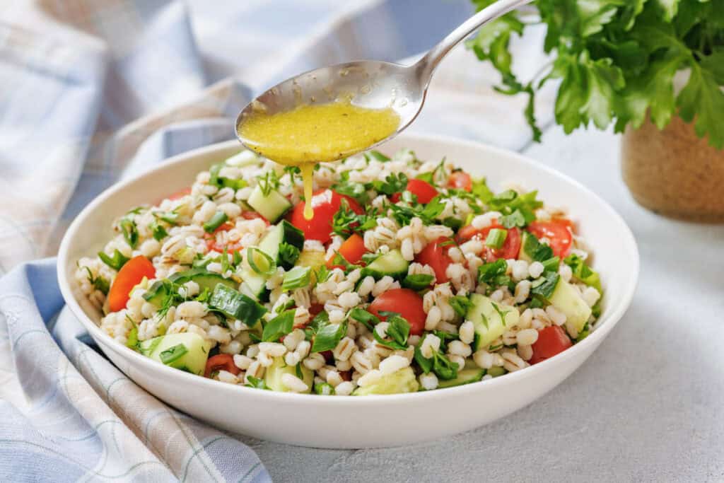 A spoon drizzles dressing over a bowl of barley salad with chopped cucumbers, tomatoes, and herbs on a light tablecloth.