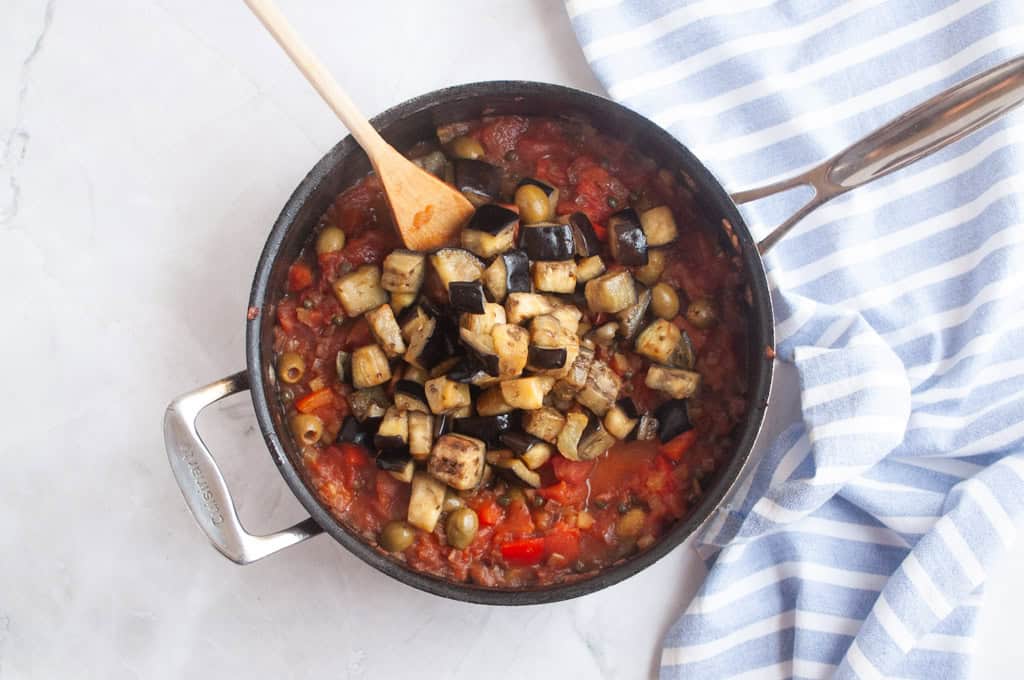 A pan of vegetable stew with diced eggplant, olives, and tomato sauce sits on a marble surface next to a blue-and-white striped cloth. A wooden spoon rests inside the pan.