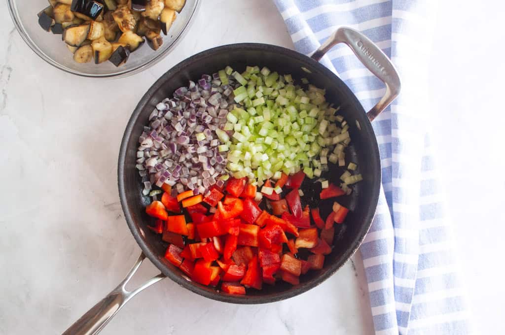 A frying pan with chopped red bell pepper, celery, and red onion arranged separately; a bowl of cooked eggplant and a striped towel are nearby.