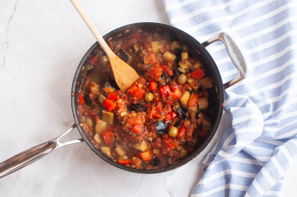 A pan of vegetable stew with tomatoes, eggplant, olives, and peppers sits on a marble surface next to a blue and white striped towel. A wooden spoon rests in the pan.