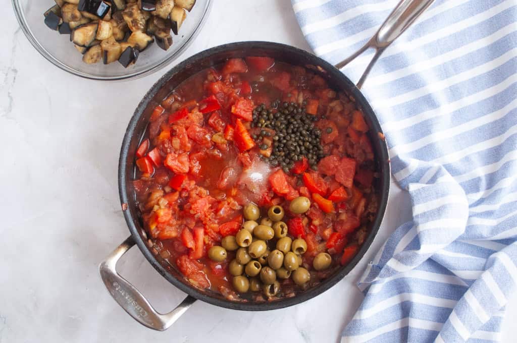 A pan filled with tomato sauce, chopped tomatoes, green olives, and capers sits on a marble surface next to a bowl of cooked eggplant and a blue-striped towel.