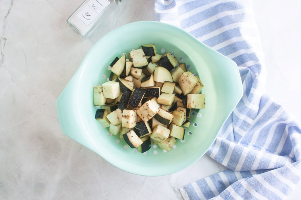 Cubed eggplant in a mint green colander on a marble surface, with a striped blue dish towel and a salt container nearby.