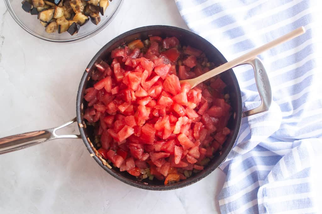 A pan filled with diced tomatoes and vegetables sits on a white surface beside a blue-and-white striped towel and a bowl of cooked eggplant.
