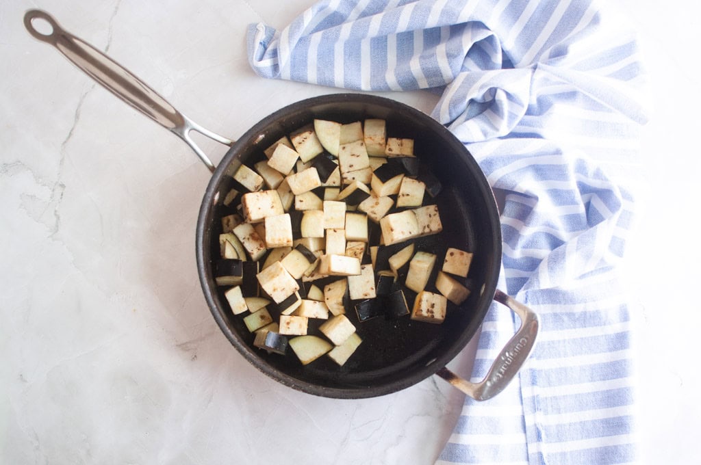 A black pan with diced eggplant pieces sits on a light countertop next to a blue and white striped kitchen towel.