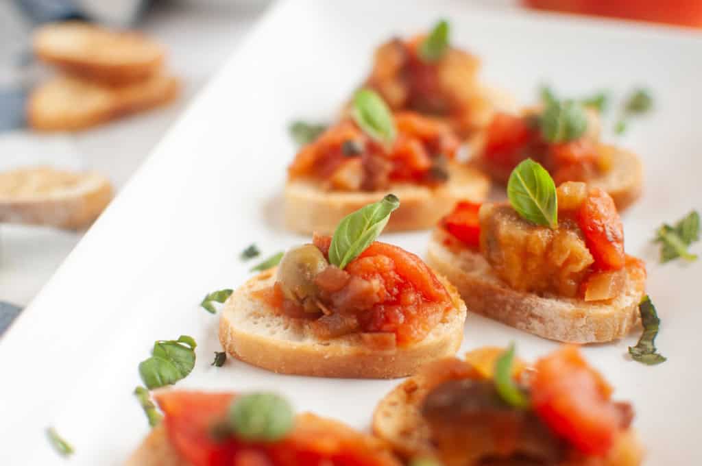 Small slices of toasted bread topped with a tomato-based vegetable mixture and garnished with fresh basil leaves, arranged on a white plate.
