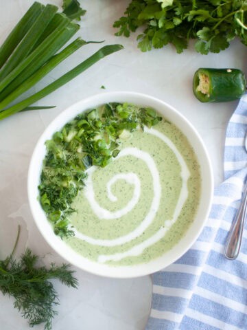 Overhead shot of a bowl of creamy cucumber soup with herbs and a swirl of cream, alongside a spoon, cloth, and vegetables.