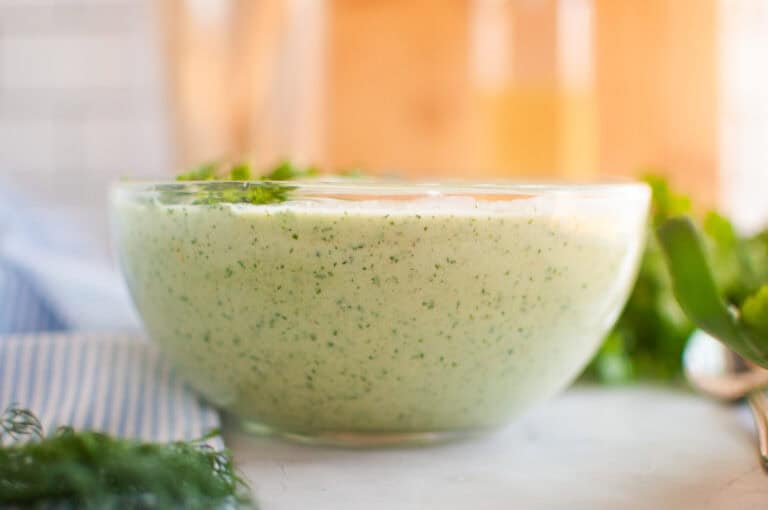 A clear glass bowl filled with creamy cucumber soup, garnished with herbs.