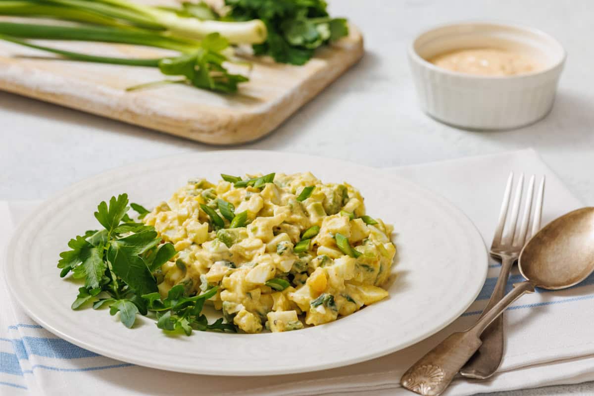 A white plate with avocado egg salad, garnished with herbs, is shown with a fork, spoon, cloth, and a small bowl of sauce.