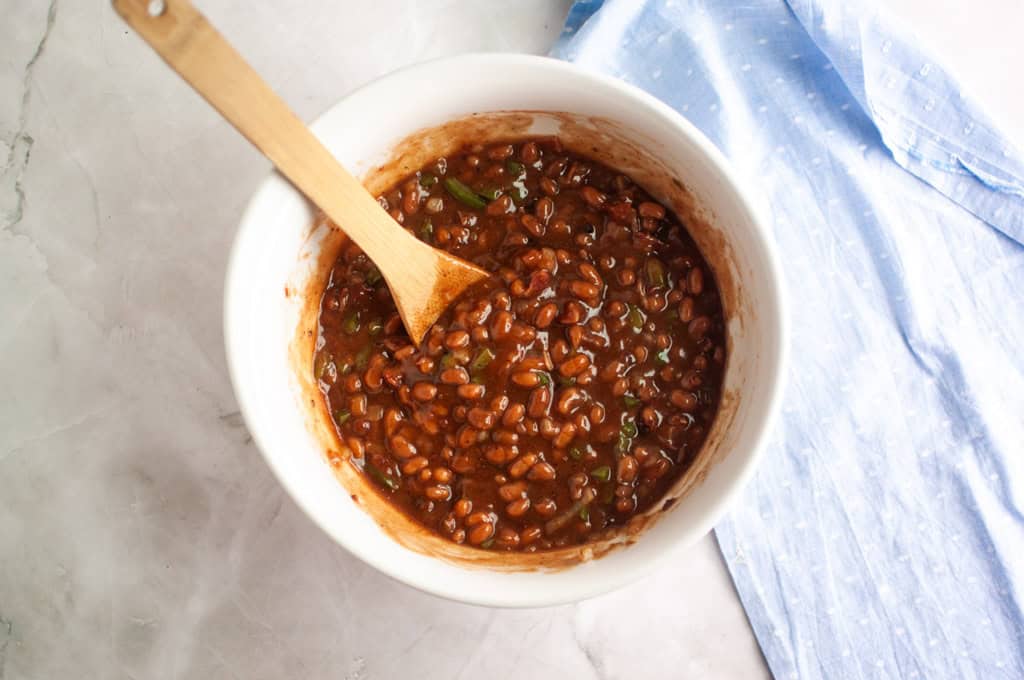 A white bowl filled with a mixture of baked beans and green peppers, stirred with a wooden spoon, sits on a light marble surface next to a blue cloth.