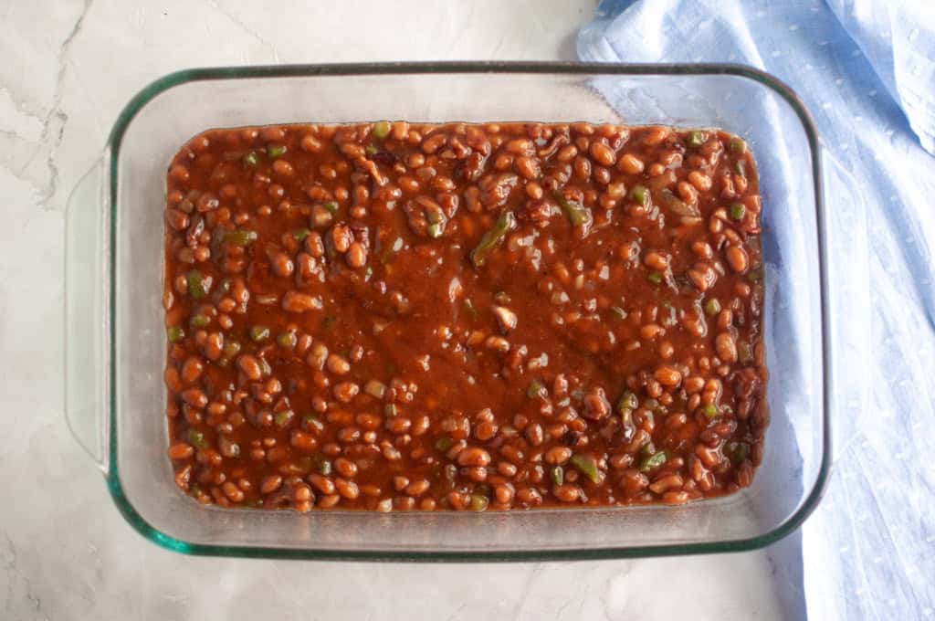 A glass baking dish filled with baked beans in sauce on a light surface, with a blue cloth partially visible to the side.