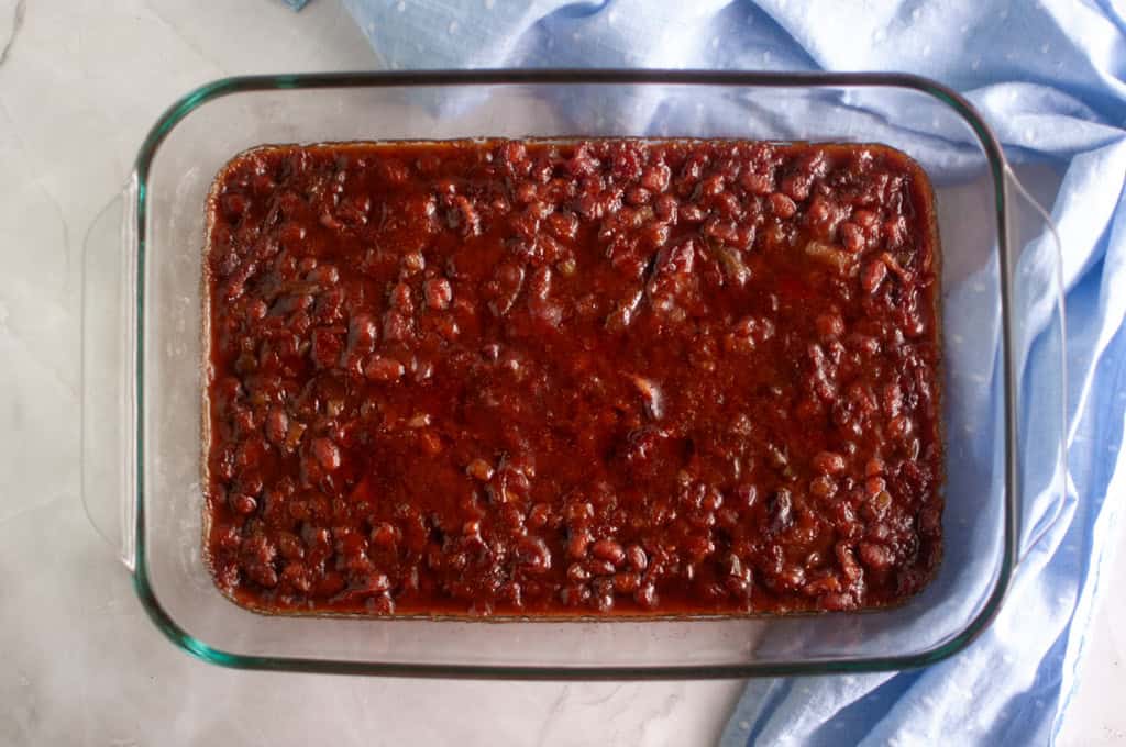 A glass baking dish filled with baked beans in a thick, reddish-brown sauce, placed on a light countertop next to a blue cloth.