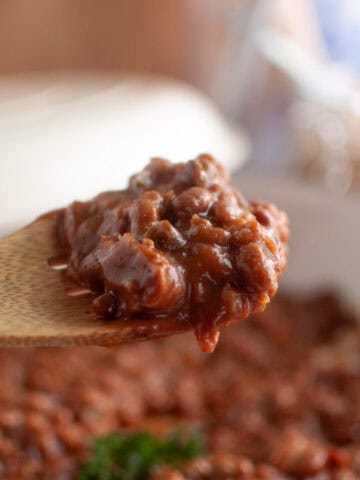 A wooden spoon holds a serving of Baked Bean Casserole above a pot filled with more bean casserole.