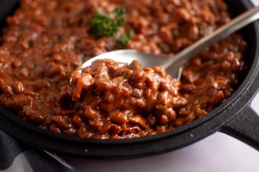A close-up of baked beans in a cast iron skillet, with a spoon scooping some of the beans and a parsley garnish in the background.