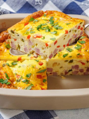 A hand holds a spatula lifting one piece of Grandma’s Baked Western Omelet from a square dish.