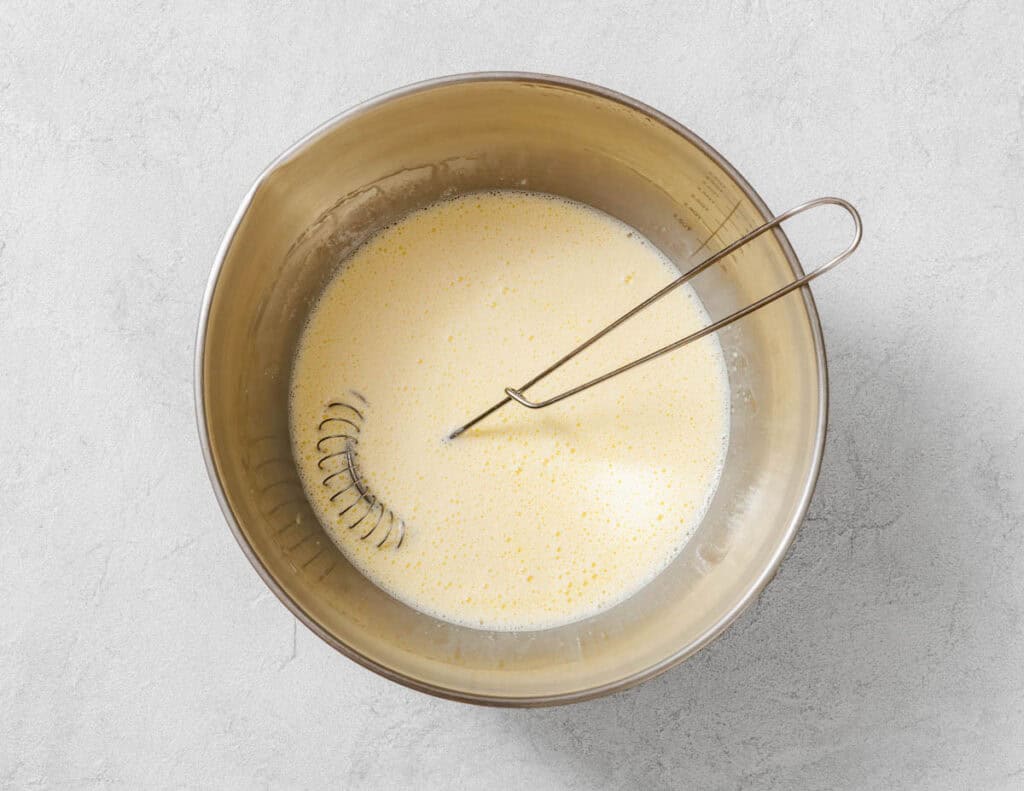 A metal mixing bowl filled with a pale yellow liquid mixture and a metal whisk resting inside, on a light gray surface.