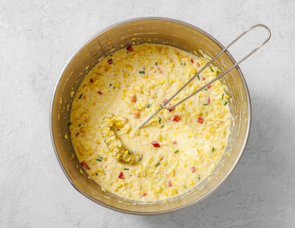 A metal bowl containing a creamy batter with chopped vegetables and corn, being mixed with a whisk on a light gray surface.