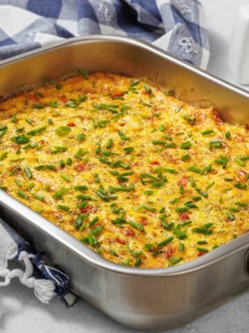 A Corn Pudding Casserole with chopped chives on top is shown in a stainless steel dish on a table with a checkered cloth and salt and pepper shakers nearby.