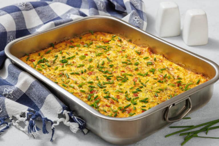A corn pudding casserole with chopped chives on top is shown in a stainless steel dish on a table with a checkered cloth and salt and pepper shakers nearby.