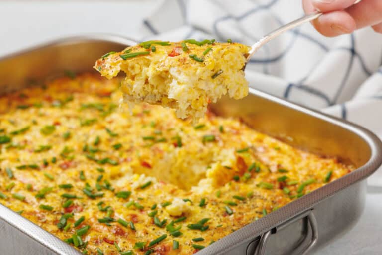 A corn pudding casserole topped with chopped chives in a metal baking dish, with a portion being lifted out on a serving spatula.