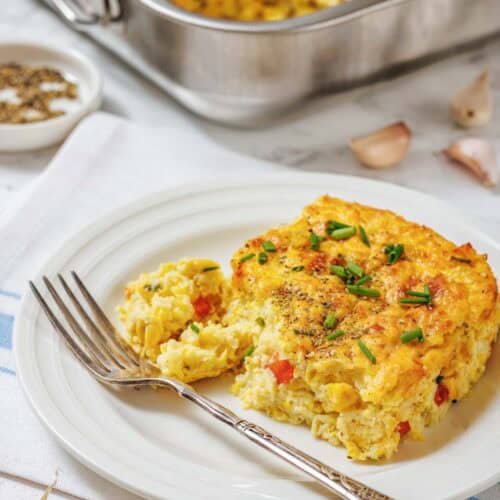 A slice of Corn Pudding Casserole with vegetables is served on a white plate with a fork. The casserole dish and seasoning are visible in the background.