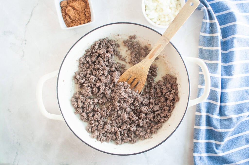 Overhead shot of cooked ground beef in a white pot with a wooden spoon, with bowls of spices and onions nearby.