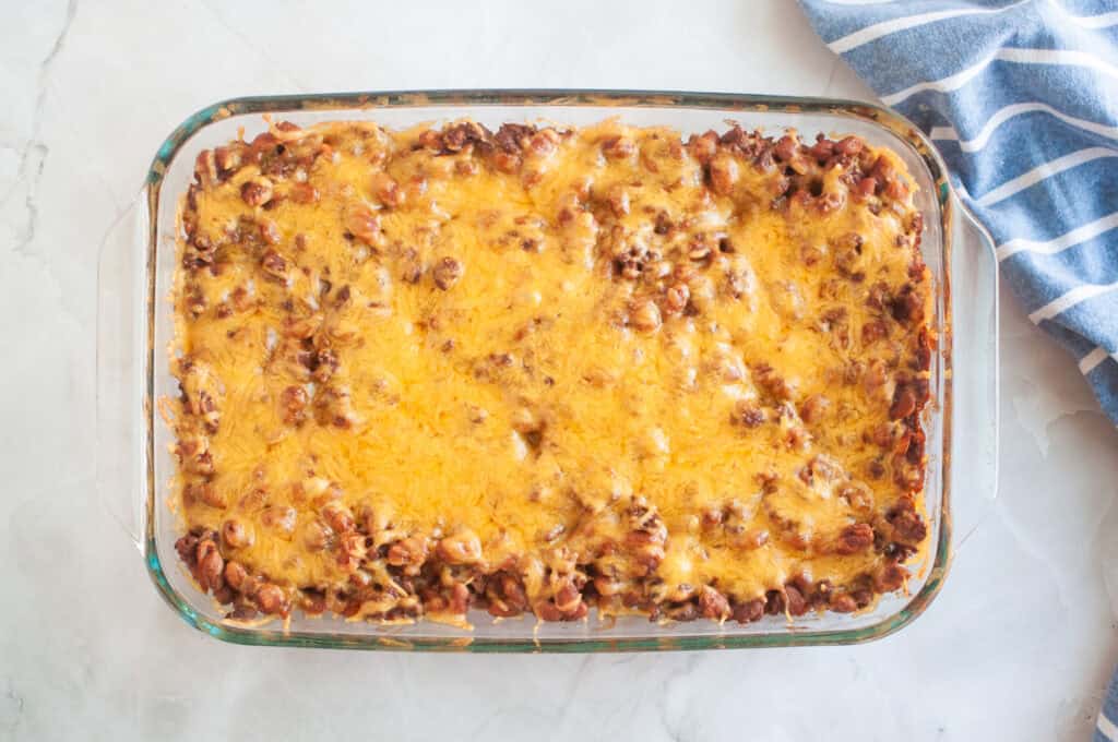 Overhead shot of a baked casserole with beans, ground beef, and melted cheese in a clear glass dish, with a blue striped cloth beside it.