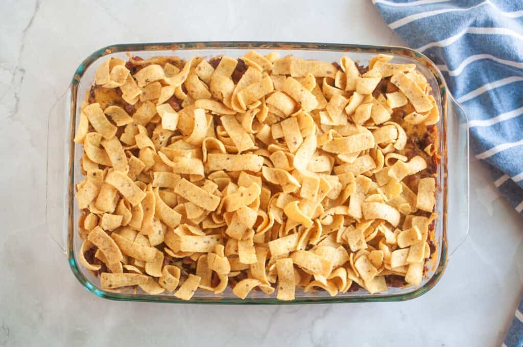 Overhead shot of a casserole with beans, ground beef, cheese, and chips in a clear glass baking dish, with a blue striped cloth beside it.