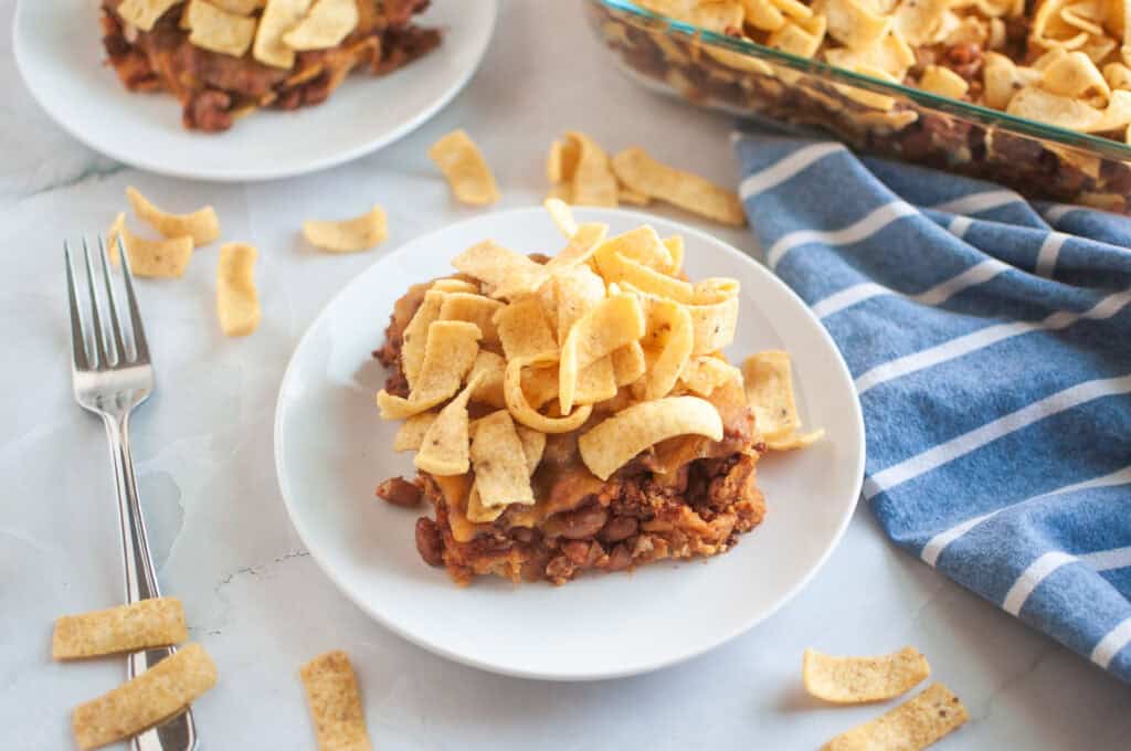 A white plate with a serving of frito pie, topped with corn chips, is shown on a light surface. A fork, a baking dish with more pie, and a blue striped cloth are visible.