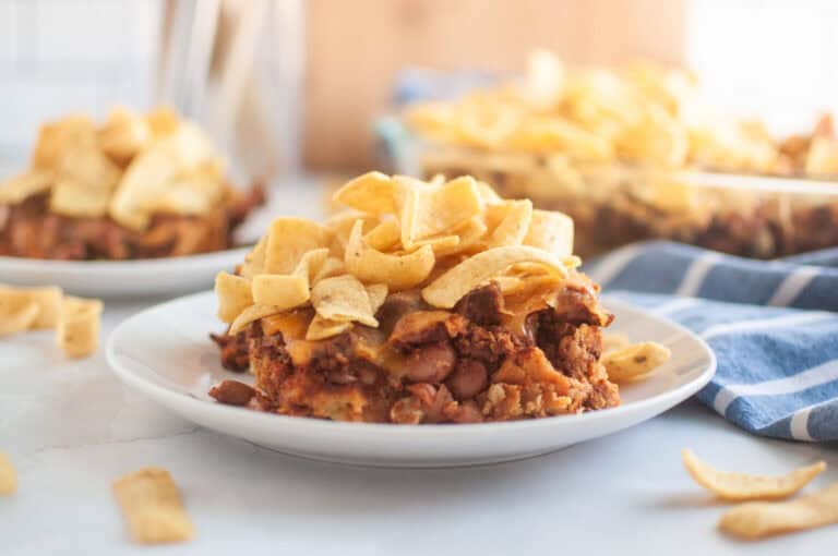 A serving of Frito pie on a white plate, topped with corn chips and melted cheese, with more Frito pie and chips in the background.