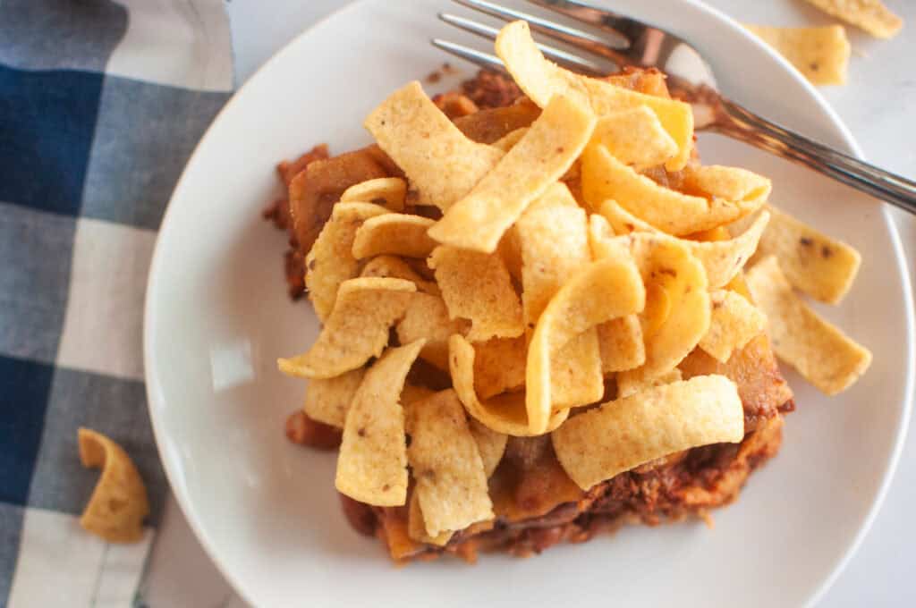 A white plate with a serving of frito pie, topped with corn chips, is shown with a fork. A blue checkered cloth is visible in the background.