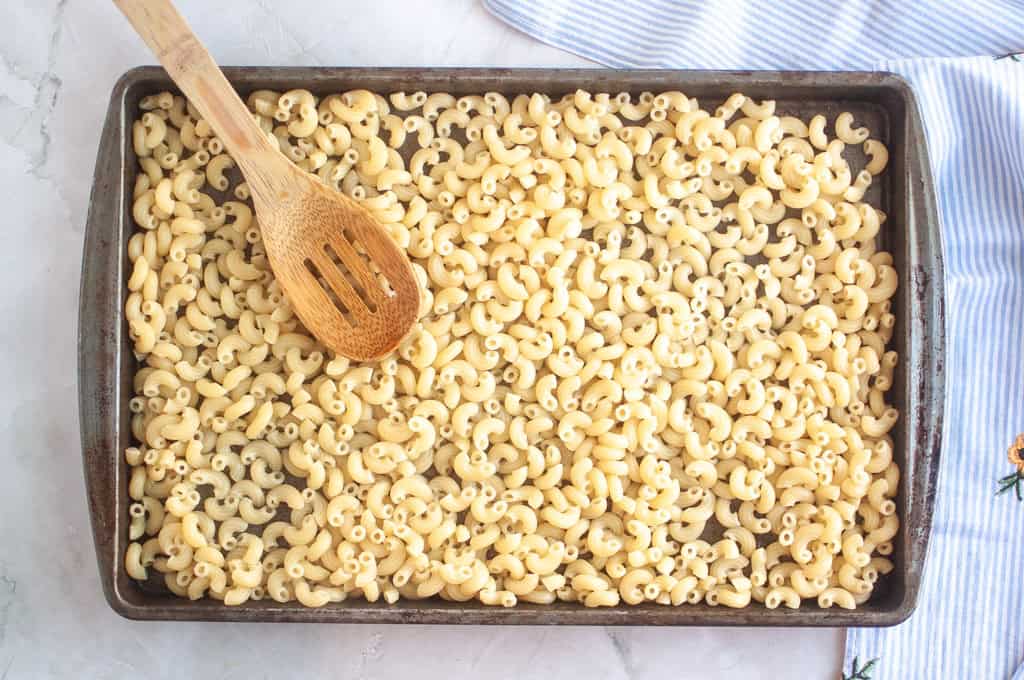 Overhead shot of cooked elbow macaroni spread on a baking sheet with a wooden spoon, on a light surface with a striped cloth.