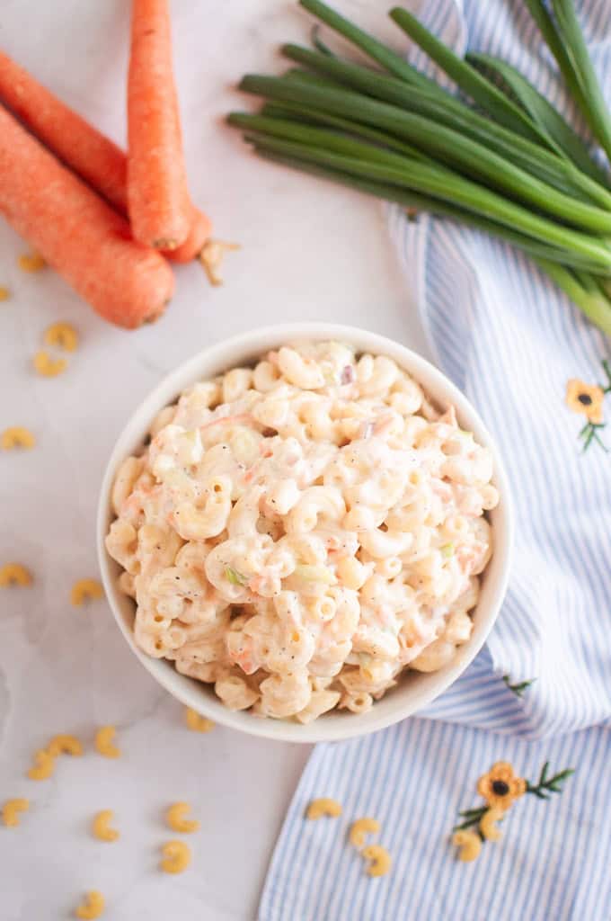A white bowl filled with macaroni salad, containing elbow macaroni and a creamy dressing, is shown with carrots and scallions on the side.