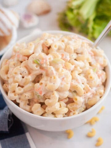A white bowl filled with macaroni salad, containing elbow macaroni and a creamy dressing, is shown with a spoon inside. Garlic cloves and lettuce are visible around the bowl.