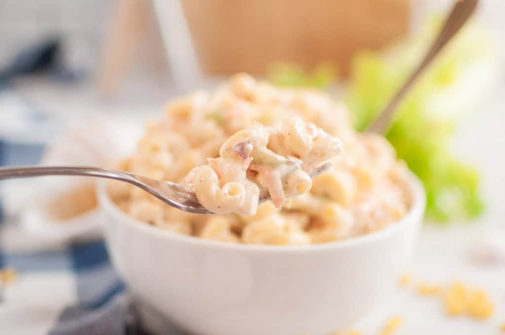 A close-up shot of a fork lifting a portion of macaroni salad, with a bowl of the salad in the blurred background.