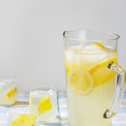 A refreshing glass pitcher filled with homemade lemonade, ice cubes, and fresh lemon slices, accompanied by three glasses of lemonade on a plaid tablecloth.