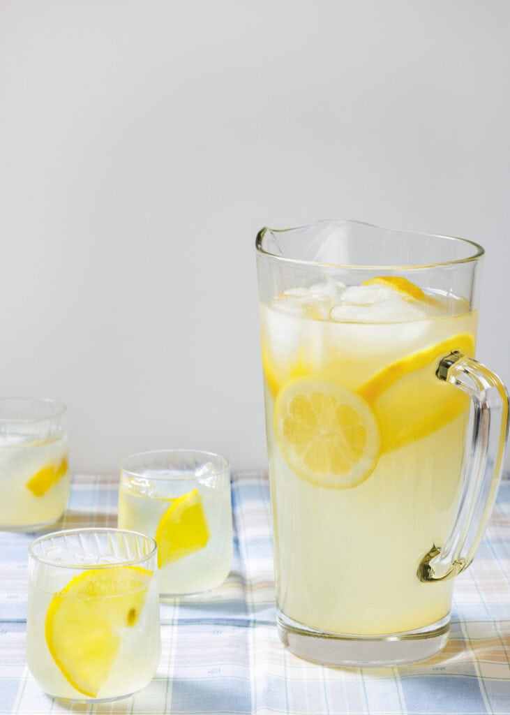 A refreshing glass pitcher filled with homemade lemonade, ice cubes, and fresh lemon slices, accompanied by three glasses of lemonade on a plaid tablecloth.