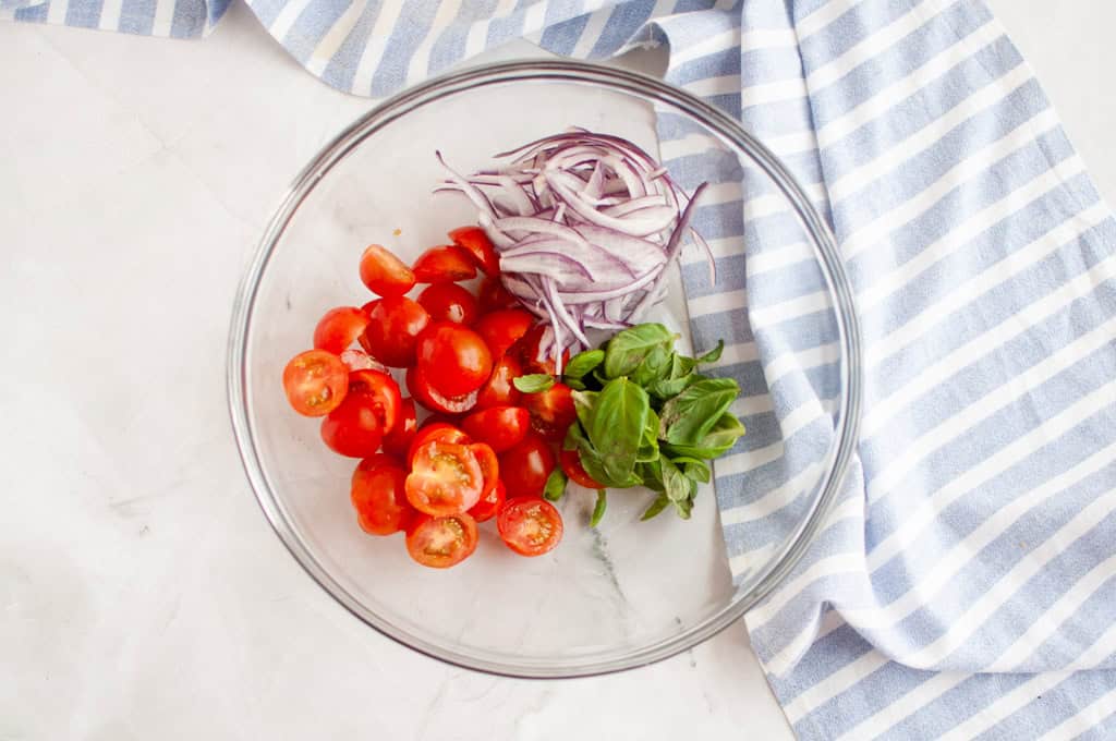 Overhead shot of cherry tomatoes, red onion, and basil in a glass bowl, with a striped cloth.